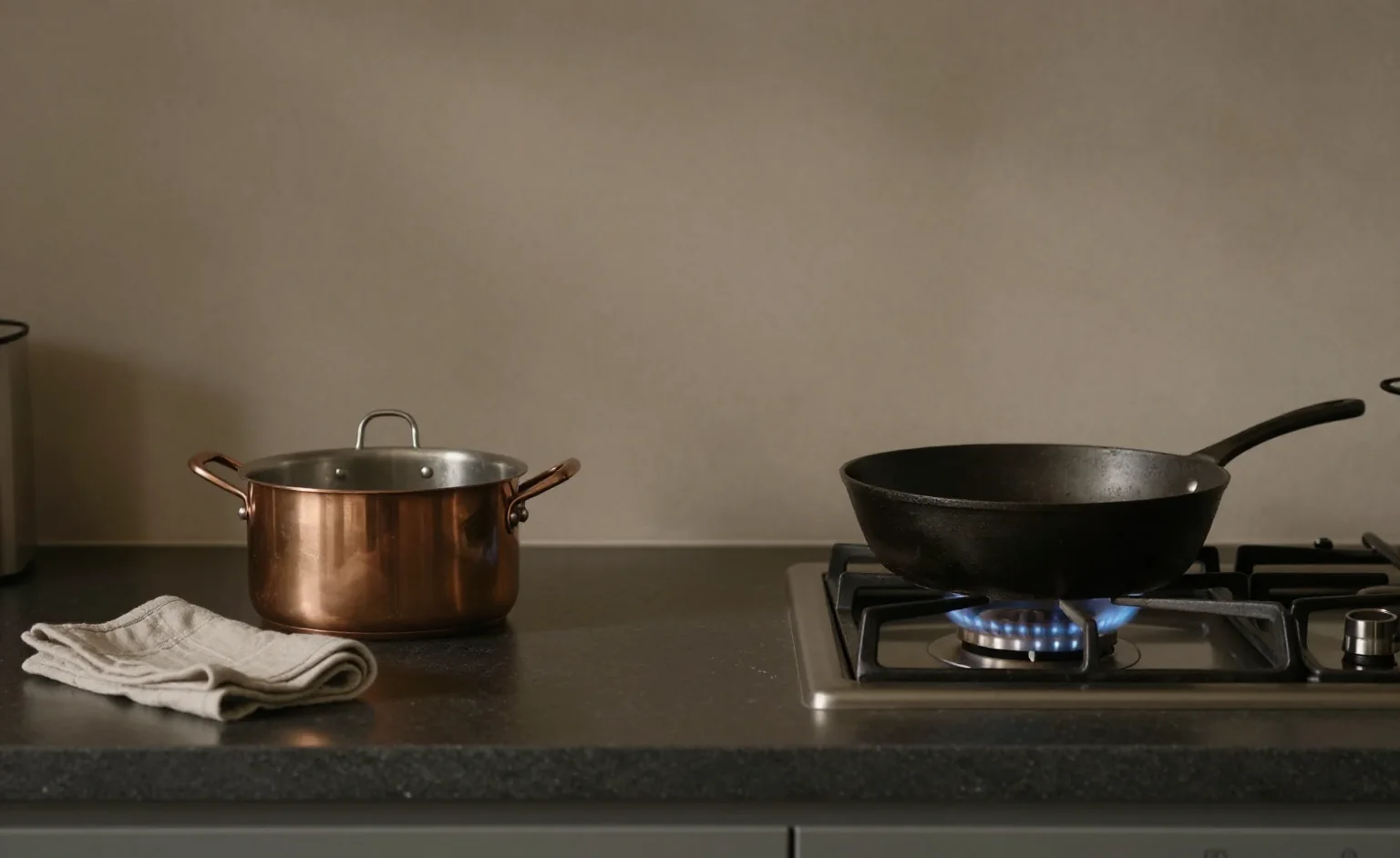 A feng shui kitchen counter at dusk: a copper pot of water and a folded cloth on the left, a lit gas hob with a cast-iron pan on the right, and clear dark worktop between them